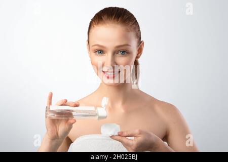 Hautpflege. Lächelnd Rothaarige Teen Girl Anwendung Lotion Auf Cotton Pad Reinigung Der Haut Entfernen Make-Up Posiert Über Grauen Hintergrund. Studioaufnahme. Natürlich Stockfoto