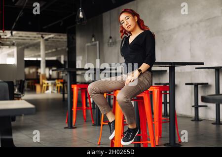 Schöne modische asiatische Frau in Brille posiert lächelnd Blick auf die Kamera, glückliche asiatische Frau lächelnd und posiert in einem Loft-Café auf einer BA sitzen Stockfoto