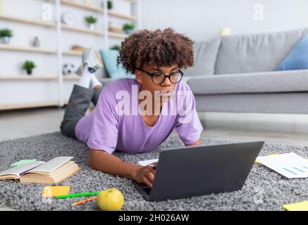 Fokussierter schwarzer Teenager, der Laptop-pc für Fernunterricht verwendet, auf dem Boden liegt und zu Hause auf der Tastatur tippt. Afroamerikanischer Jugendlicher, der teilnimmt Stockfoto