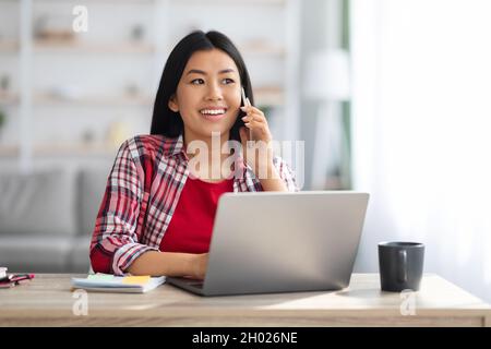 Home Office-Konzept. Junge Asiatische Frau Im Gespräch Auf Handy Und Mit Laptop, Fröhliche Koreanische Freiberuflerin Dame Remote Mit Computer Und Enjo Arbeiten Stockfoto