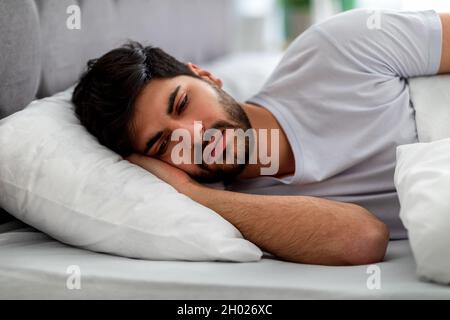 Trauriger arabischer Kerl, der allein im Bett liegt, unter Einsamkeit leidet und an Probleme denkt. Männlicher Stress und Depression, Schlaflosigkeit und Apathie Konzept. Stockfoto