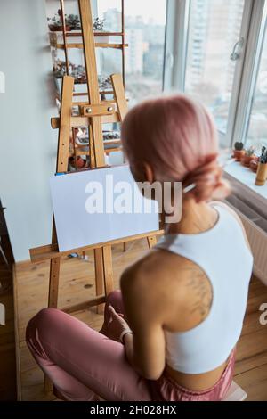 Frau mit Brötchen auf dem Kopf sitzt an der Staffelei auf dem hellen Balkon zu Hause Stockfoto