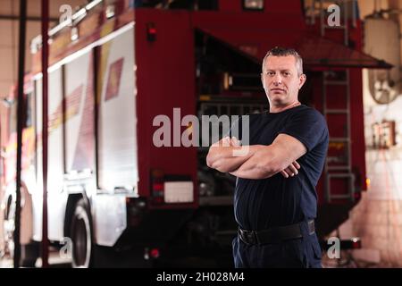 Professionelles Feuerwehrporträt. Feuerwehrmann trägt Uniform aus Hemd und Hose. Feuerwehrauto im Hintergrund. Stockfoto