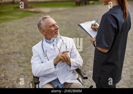 Lächelnder leitender, behinderter Arzt im Rollstuhl mit Laborkittel, der sich mit der Krankenschwester unterhält, die sich Notizen macht, während er ihm im Freien hilft Stockfoto