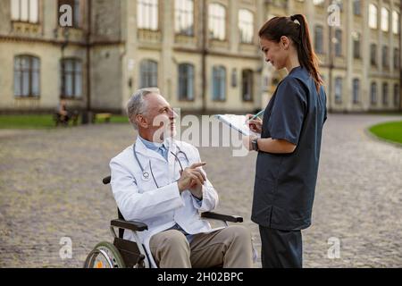 Erfolgreicher älterer, behinderter Arzt im Rollstuhl, der einen Laborkittel trägt und sich mit der jungen Krankenschwester unterhält, die sich Notizen macht, während er ihm im Freien hilft Stockfoto