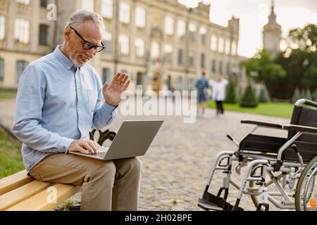 Älterer Mann, der einen Patienten mit Rollstuhl wiederfindet, der einen Videoanruf abruft, während er auf der Bank im Park in der Nähe der Klinik sitzt und an einem Laptop arbeitet Stockfoto