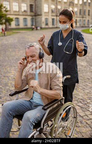 Junge Krankenschwester in Schutzmaske, die sich im Rollstuhl um einen älteren Patienten kümmert, der Kopfhörer trägt und im Park Musik hört Stockfoto