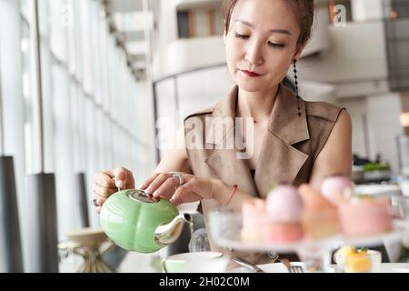 Reife asiatische Frau, die im Café in der Hotellobby am Tisch sitzt und Dessert isst und Tee trinkt Stockfoto