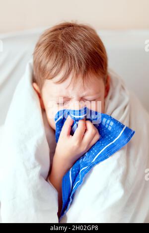 Der Junge bläst seine Nase in ein Taschentuch, eingewickelt in eine Decke. Krankes Kind. Eine Erkältung mit einer laufenden Nase in einem Jungen Stockfoto