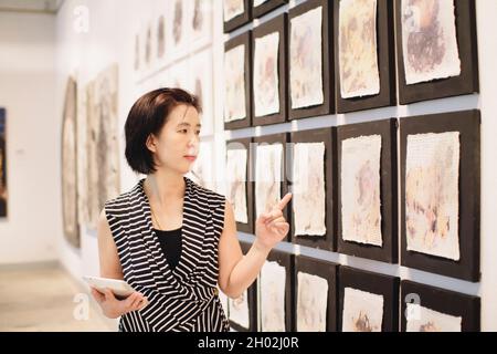 Asiatische Frau, die in einer Kunstgalerie vor farbenfroh gerahmten Gemälden an einer Wand steht Stockfoto