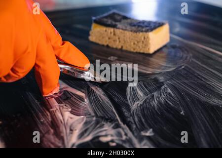 Nahaufnahme eines orangefarbenen Handschuhs mit einem Metallschaber, der das Kochfeld reinigt. Selektiver Fokus. Ein gelber Schwamm liegt im Hintergrund. Das Konzept von Home cl Stockfoto