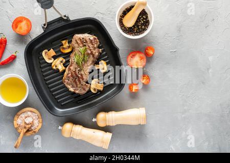 Gegrilltes Rindersteak mit Pilzen und Gewürzen auf einer Grillpfanne auf grauem Hintergrund Stockfoto