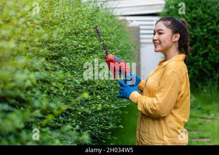 Junge Frau, die zu Hause im Garten eine kabellose elektrische Heckenschneide- und Schneideanlage verwendet Stockfoto