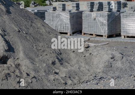 Bauindustrie, Baustoffe. Paletten aus grauen Pflasterfliesen und einem Sandhaufen. Neue Zementblöcke und Bordsteine in Plastikfolie verpackt. Keine pe Stockfoto