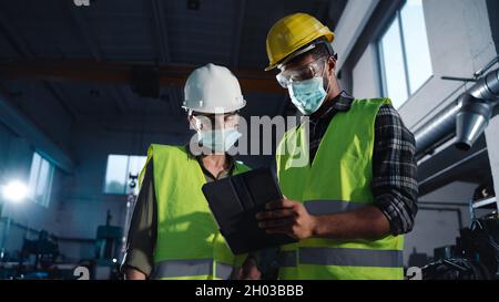 Low-Angle-Ansicht von Industrieinspektoren mit Gesichtsmasken, die eine allgemeine Überprüfung in Innenräumen in Metallwerkstatt durchführen, Coronavirus-Konzept. Stockfoto