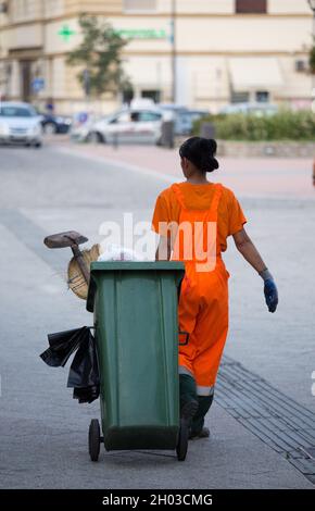 Rückansicht einer Müllarbeiterin in orangefarbener Uniform, die den Mülleimer mit anderen Geräten zieht Stockfoto