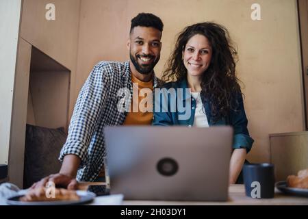 Glückliches biracial Paar mit Laptop sitzen, ruhen und betrachten Kamera drinnen in einem Baumhaus, Wochenende weg und Remote-Office-Konzept. Stockfoto