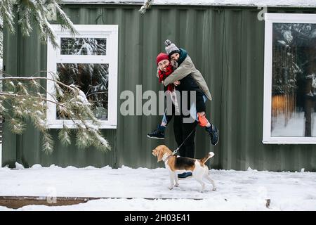 Verspieltes Paar posiert für ein Foto, der Mann trägt die Frau auf dem Rücken. Türen im Winter. Vor dem Wellblechhaus. Stockfoto