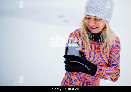 Aktive ältere Läuferin, die das Smartphone am Arm anpasst, um im Schnee im Freien zu trainieren. Stockfoto