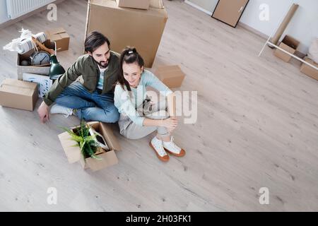Blick aus der Ferne auf ein junges Paar mit auf dem Boden sitzenden Boxen beim Umzug in eine neue Wohnung, ein neues Zuhause und ein Umzugskonzept. Stockfoto