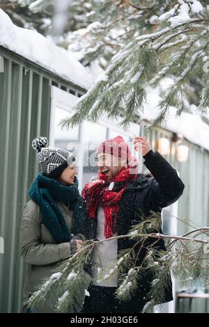 Fröhliches Paar, das sich unterhielt und von den Bäumen in der Nähe Schnee über sich hereinschneite. Beide tragen Hüte, Jacken und Schals. Vor einem Haus mit Corrugate Stockfoto