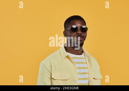 Minimal Waist up Portrait des trendigen afroamerikanischen Mannes mit Sonnenbrille und Blick auf die Kamera, während er vor gelbem Hintergrund im Studio steht, Kopierraum Stockfoto