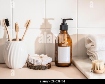 Mockup für Pflegekosmetikprodukte - Bernsteinflasche Hautpflege mit weißen Etiketten, Bambuszahnbürste und verschiedenem Zubehör auf beigem Tisch und Wand weiß Stockfoto