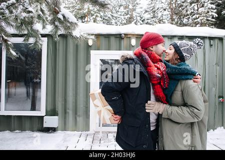 Selbstbewusster Mann in Winterkleidung küsst seine Frau, während er die Gegenwart hinter seinem Rücken versteckt. Vor einem kleinen verschneiten Haus stehen. Stockfoto