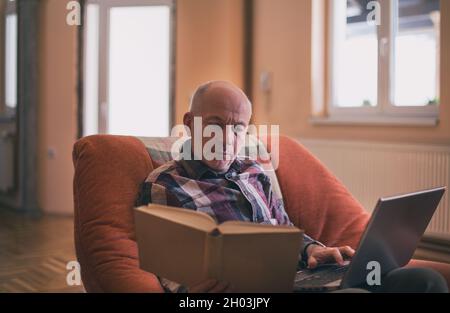 Reifer Mann, der im Sessel sitzt und am Laptop und im Buch recherchiert Stockfoto