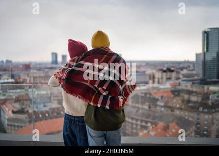 Rückansicht eines jungen Paares, das in eine Decke gehüllt und auf dem Balkon im Freien steht und sich umarmt, mit Blick auf die Stadt. Stockfoto