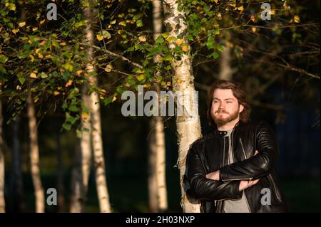 Ein junger bärtiger Mann, der im Herbstpark bei den Bäumen steht und nachdenklich in die Ferne blickt Stockfoto