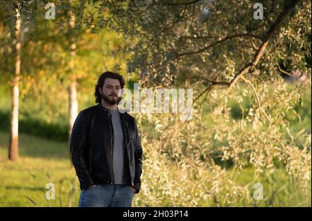 Ein junger bärtiger Mann, der im Herbstpark bei den Bäumen steht und nachdenklich auf die Kamera schaut Stockfoto