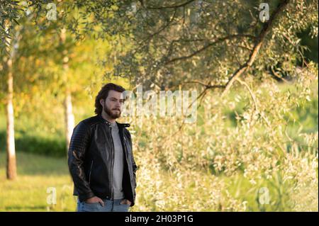 Ein junger bärtiger Mann, der im Herbstpark bei den Bäumen steht und nachdenklich in die Ferne blickt Stockfoto