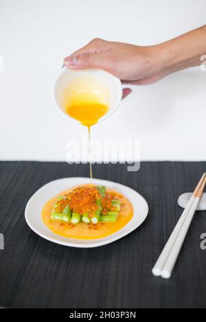 Gießt Sauce von Hand auf Minispargel mit rotem Kaviar. Weißer Teller und Essstäbchen auf schwarzer Matte im japanischen Restaurant, Nahaufnahme, Bild für die Speisekarte Stockfoto