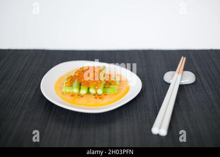 Mini-Spargel mit Sauce und rotem Kaviar auf weißem Teller mit Essstäbchen auf schwarzer Matte im japanischen Restaurant, Nahaufnahme, flaches Lay, Bild für die Speisekarte Stockfoto