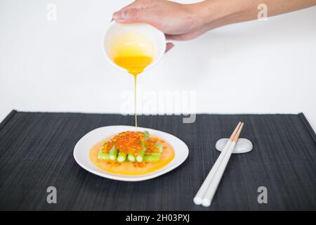 Gießt Sauce von Hand auf Minispargel mit rotem Kaviar. Weißer Teller und Essstäbchen auf schwarzer Matte im japanischen Restaurant, Nahaufnahme, Bild für die Speisekarte Stockfoto