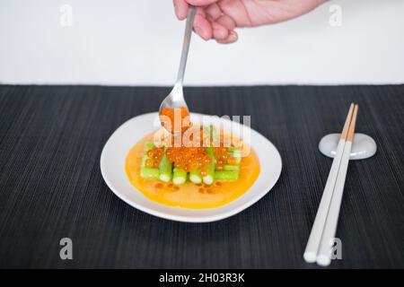 Hand legt roten Lachs-Kaviar auf Mini-Spargel mit Sauce auf weißem Teller im japanischen Restaurant, Nahaufnahme, flaches Lay, Bild für die Speisekarte, gesundes Essen Stockfoto