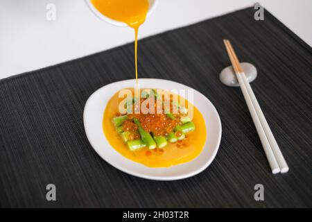 Gießt Sauce von Hand auf Minispargel mit rotem Kaviar. Weißer Teller und Essstäbchen auf schwarzer Matte im japanischen Restaurant, Nahaufnahme, Bild für die Speisekarte Stockfoto