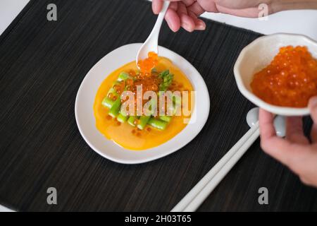 Hand legt roten Lachs-Kaviar auf Mini-Spargel mit Sauce auf weißem Teller im japanischen Restaurant, Nahaufnahme, flaches Lay, Bild für die Speisekarte, gesundes Essen Stockfoto