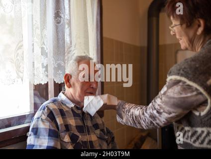 Reife Frau hilft älteren Mann, Nase zu blasen, indem sie Serviette hält Stockfoto