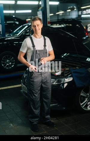 In voller Länge vertikale Porträt von lächelnden schönen jungen Mechaniker männlich trägt Uniform mit speziellen Schlüssel Ratschenschlüssel. Stockfoto