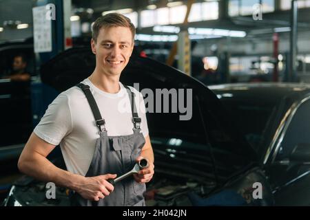 Mittellange Aufnahme eines fröhlichen, hübschen jungen Mechanikers mit Uniform und speziellem Schlüsselschlüssel Stockfoto