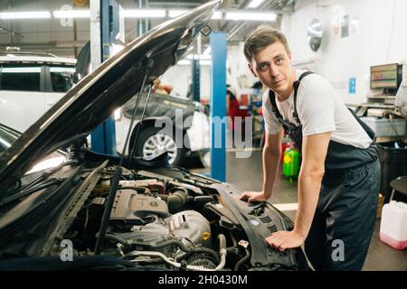 Seitenansicht des selbstbewussten, gutaussehenden professionellen Kfz-Mechanikers in blauer Uniform, der vor der offenen Haube steht. Stockfoto