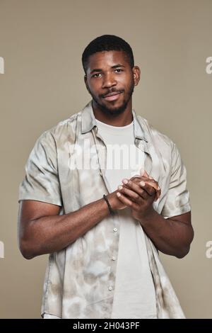 Vertikales Porträt eines hübschen afroamerikanischen Mannes, der Hemd trägt und die Kamera anschaut, während er im Studio vor neutralem beigen Hintergrund posiert Stockfoto
