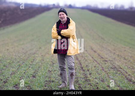 Portrait eines älteren Bauern, der im Winter gekreuzte Arme in einem Weizenfeld mit kleinen Pflanzen hält Stockfoto