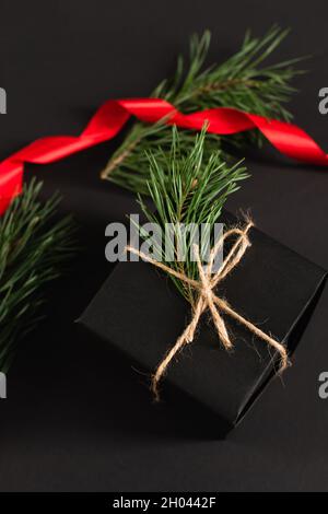 Draufsicht auf Geschenkbox mit Tannenzweig in der Nähe von verschwommenem rotem Band auf schwarz Stockfoto