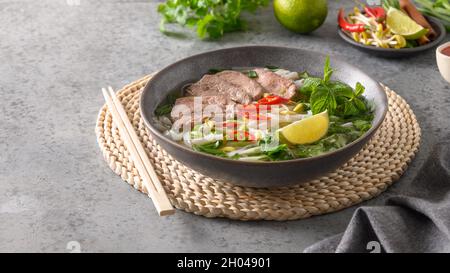 Pho Bo Suppe mit Rindfleisch in grauer Schale auf grauem Steingrund. Nahaufnahme. Vietnamesische und asiatische Küche. Stockfoto