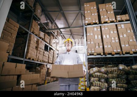 Lächelnde Arbeiterin, die Kisten im Lager trägt. Schutzhelm am Kopf, rund um Boxen auf Regalen. Stockfoto