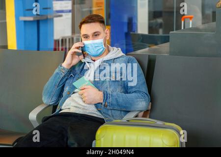 Attraktiver junger Mann in medizinischer Gesichtsmaske, der mit dem Smartphone in der Flughafenlounge mit dem Koffer redet, den Reisepass vor der Reise in der Hand hält Stockfoto