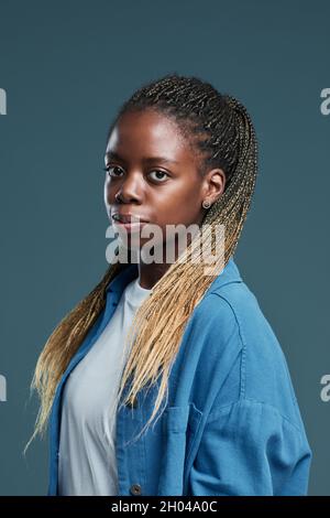 Vertikales Porträt einer modernen afroamerikanischen jungen Frau mit Afro-Zöpfen-Frisur, die vor blauem Hintergrund auf die Kamera blickt Stockfoto